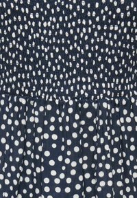 Donkerblauwe stof met witte stippen; het bovenste gedeelte is strak geplooid of gesmokt, het onderste gedeelte is los en vloeiend.