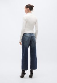 Εφαρμοστή λευκή μπλούζα με μακριά μανίκια, συνδυασμένη με ψηλόμεσα, φαρδιά μπλε τζιν παντελόνια. Τα τζιν έχουν δύο πίσω τσέπες και εφέ ξεθωριάσματος.