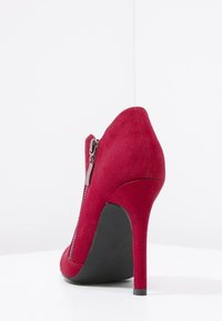 Chaussure à talon haut en daim rouge avec une silhouette élégante, dotée d'une fermeture éclair latérale et d'un bout pointu. Semelle en caoutchouc noire pour une meilleure adhérence.