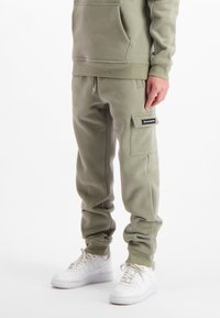 Persoon in olijfgroene cargojoggingbroek met een zijzak en witte sneakers, staand tegen een witte achtergrond.