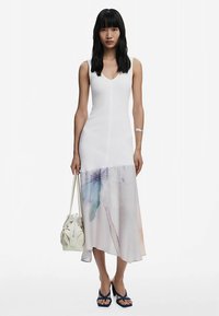Robe blanche sans manches avec un col en V, présentant un imprimé en dégradé de couleurs pastel douces à l'ourlet. Le mannequin porte un sac à main texturé.