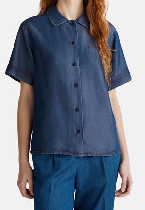 Mujer con una camisa vaquera de manga corta abotonada y pantalones vaqueros azules a juego sobre un fondo liso.