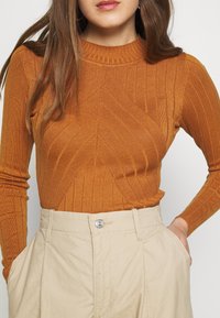 Top orange à col roulé côtelé avec un motif chevron texturé, associé à un pantalon beige taille haute avec un bouton à l'avant et des poches latérales.