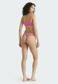 Frau steht barfuß, mit dem Rücken zur Kamera, trägt einen pinkfarbenen Sport-BH und passende String-Unterwäsche vor einem schlichten, hellen Hintergrund.