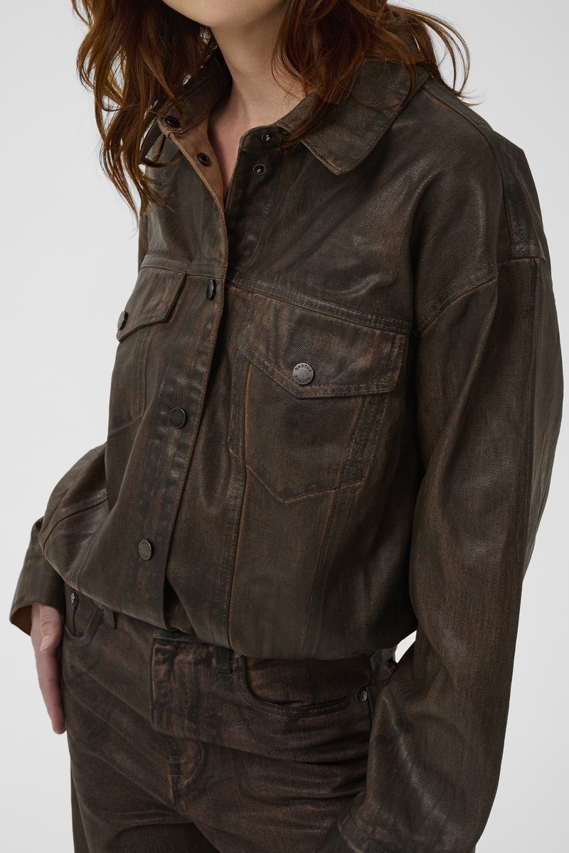 Kvinna som bär en brun sliten läderjacka med framfickor och matchande bruna byxor, stående med ena handen i fickan.