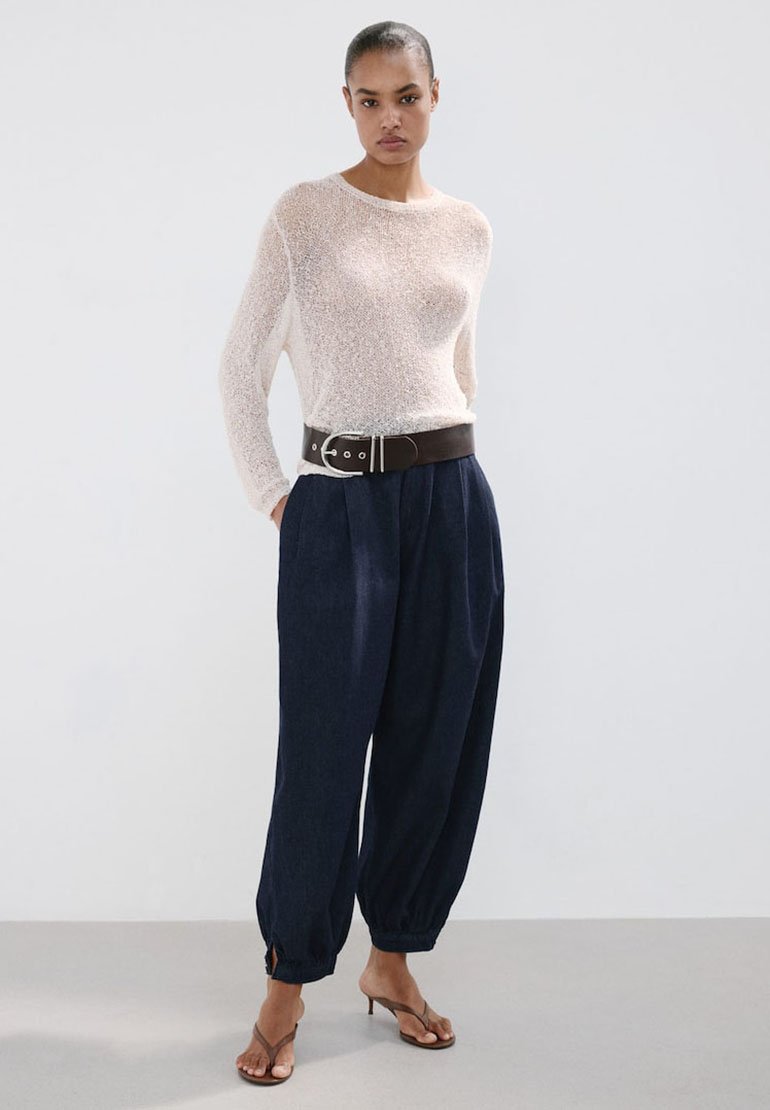 Femme portant un haut blanc transparent à manches longues, un pantalon large taille haute bleu marine avec ceinture noire, et des sandales marron à talons, debout devant un fond uni.