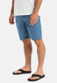 Blå shorts i mjukt tyg med en avslappnad passform och sidofickor; matchas med svarta flip-flops med platt sula och remdesig.
