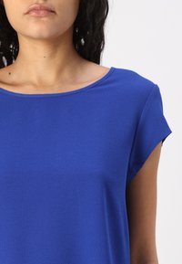 Žena v textured krátkém rukávu královsky modrém topu, ukazující krk, ramena a horní část hrudníku na čistém bílém pozadí.