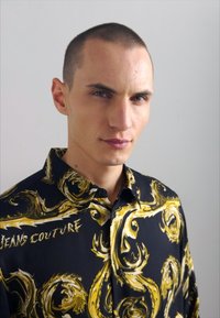 Camicia nera con audaci disegni gialli a spirale, dotata di colletto e chiusura frontale con bottoni; il tessuto sembra liscio con una finitura lucida.
