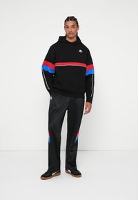 Felpa nera con strisce rosse e blu, con un logo. Abbinata a pantaloni della tuta neri, con dettagli rossi e blu. Calzature nere.
