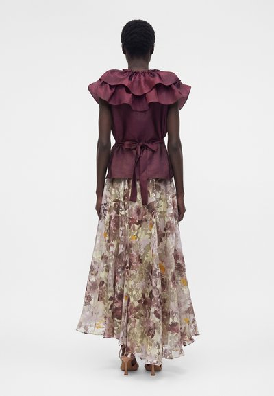 Γυναίκα που φοράει μπορντό τοπ χωρίς μανίκια με βολάν στον γιακά και ζώνη στη μέση, συνδυασμένο με μακριά, διάφανη φλοράλ φούστα και καφέ τακούνια, στέκεται γυρισμένη προς τα πίσω.