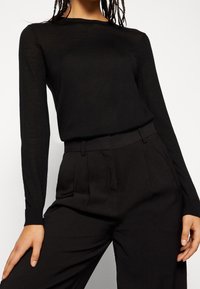 Czarny top z długim rękawem, wykonany z przezroczystego materiału, z okrągłym dekoltem i dopasowanym krojem, zestawiony z czarnymi spodniami o wysokim stanie.