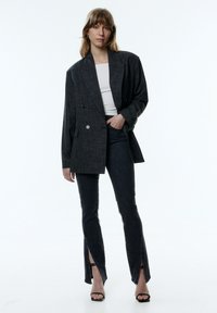 Grijze dubbele blazer met een textuurstof, gecombineerd met hoge taille jeans met voorste openingen. Zwarte gekruiste hakken maken de look compleet.