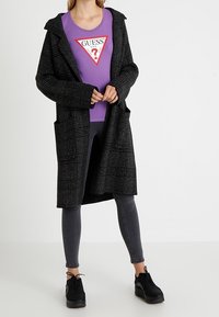 Zwarte geruite lange jas met brede mouwen en voorzakken, gedragen over een paarse t-shirt met een contrasterend logo, gecombineerd met grijze spijkerbroek en zwarte schoenen.