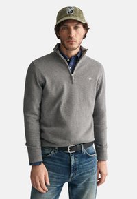 Grauer Viertelreißverschluss-Pullover aus weichem Stoff, getragen über einem blau karierten Hemd, kombiniert mit blauen Jeans und einer grünen Kappe mit dem Buchstaben 'G'.