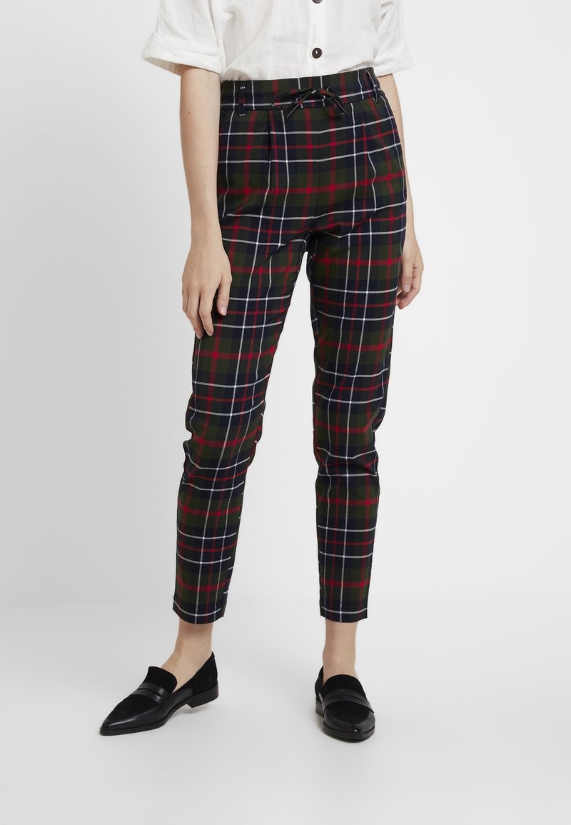 Personne portant un pantalon à carreaux taille haute, longueur cheville, en noir, rouge et blanc avec une taille nouée, associé à des mocassins noirs et une chemise blanche.