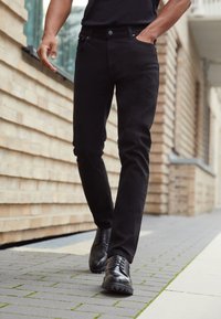 Svarta denimjeans med slim fit, femfickorsdesign och en lätt lyster, i kombination med svarta snörskor på en tegelstensväg.