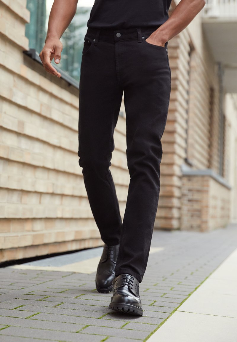 Svarta denimjeans med slim fit, femfickorsdesign och en lätt lyster, i kombination med svarta snörskor på en tegelstensväg.