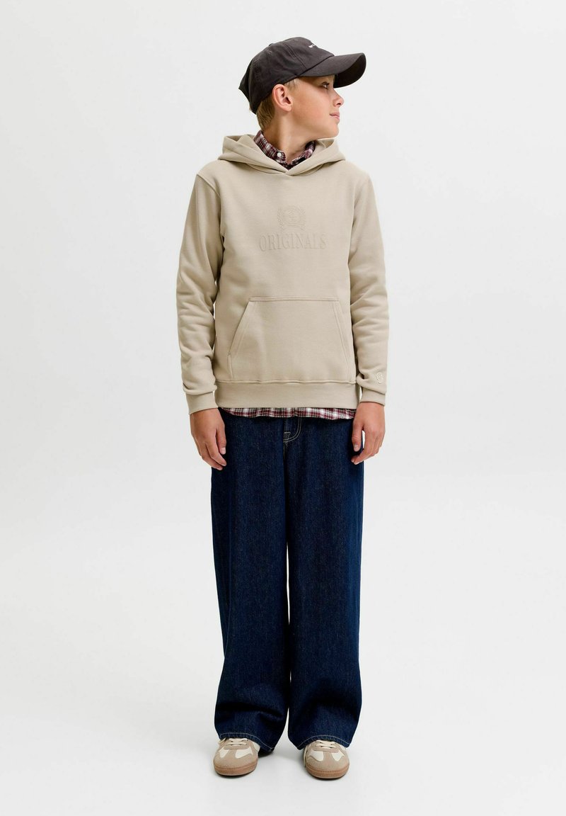 Garçon portant un sweat à capuche beige, une chemise à carreaux, un jean large foncé, des baskets beiges et une casquette de baseball noire, regardant vers sa droite sur fond blanc.