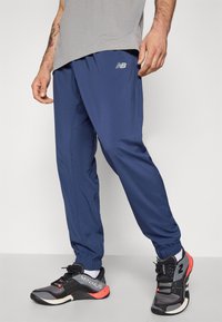 Navy blue athletic pants with elastic cuffs, featuring a small silver logo. Worn with black and red athletic shoes with white accents.