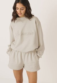 Jasno beżowy sweter z teksturowanym haftem "La mania", połączony z dopasowanymi szortami. Oba elementy charakteryzują się luźnym krojem i prążkowanym dekoltem.