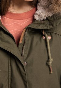 Olivgrön jacka med pälsfoder på huvan, med dragkedjestängning, beige detaljer och snörningsdetaljer. Bärs över en korallfärgad skjorta med zigzag-mönster.
