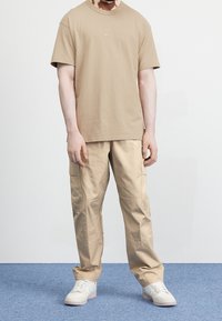 Bežové bavlněné tričko s kulatým výstřihem a malým logem, spárované s bežovými cargo kalhotami s kapsami, nošené s bílými teniskami.