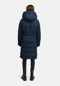 Navyblauwe pufferjas met capuchon, elastische taille en een gewatteerd ontwerp. De stof heeft een gladde textuur, en de jas reikt tot halverwege het onderbeen.