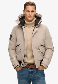 Beige pufferjakke med aftagelig hætte, sorte detaljer, to frontlommer og et logo på ærmet, lagd over en cremefarvet turtleneck.