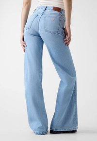 Jean évasé bleu clair avec taille haute, en denim, deux poches arrière et une étiquette en cuir de marque au-dessus de la poche droite.