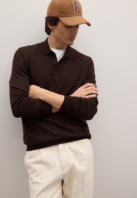 Polo in maglia marrone con colletto, abbinata a pantaloni in velluto a coste crema. Indossa un cappellino marrone con una lettera "P" nera.