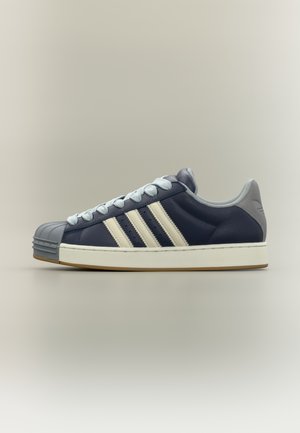 Scarpa da ginnastica in pelle blu navy con tre strisce bianche e puntale grigio texturizzato. Presenta lacci azzurri e suola in gomma. Forma arrotondata.