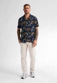 Koszula z krótkim rękawem w kolorze granatowym z motywem kwiatowym i liśćmi palmowymi, zestawiona z jasnobeżowymi spodniami i białymi trampkami. Dopasowany krój, gładka tkanina.