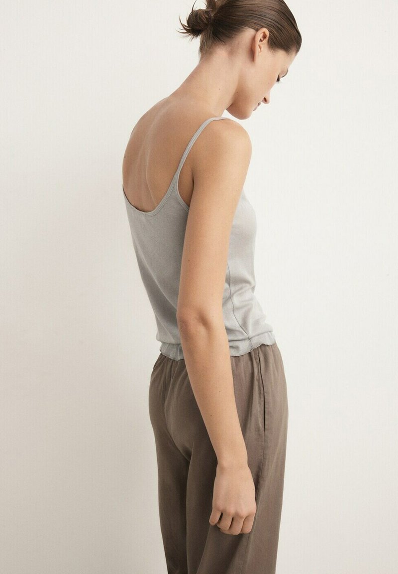 Femme aux cheveux bruns attachés en chignon, portant un haut gris clair à fines bretelles et un pantalon marron ample, debout devant un fond clair uni.