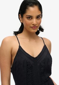 Czarna sukienka na ramiączkach spaghetti, z teksturowanej tkaniny, z dekoltem w kształcie litery V oraz skomplikowanymi koronkowymi detalami wzdłuż środka.