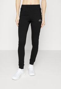 Schwarze Stretch-Jogginghosen mit schmaler Passform, ausgestattet mit seitlichen Taschen mit Reißverschluss und einem Logo-Detail an der linken Hüfte. Weiche, glatte Textur.