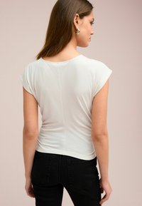 Lichtgrijs aansluitend T-shirt met korte kapmouwen en een ronde halslijn. Zachte stof met een lichte glans en geplooid detail aan de achterkant.