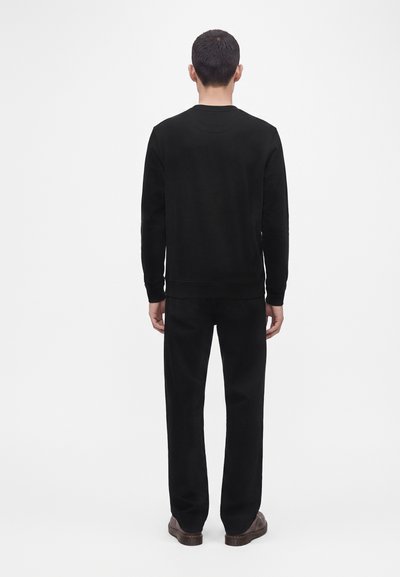 Homme aux cheveux courts et foncés portant une chemise noire unie à manches longues, un pantalon noir et des chaussures marron, debout de dos sur un fond blanc.