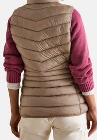 Beige Steppweste mit Zickzacknähten, getragen über einem rosa Pullover mit gerippten Manschetten. Weiße Akzente an den Manschetten. Glatte Textur.