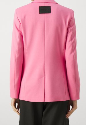 Vue de dos d'une personne portant un blazer rose vif avec une étiquette noire "HUGO" sur le haut du dos et un pantalon noir.