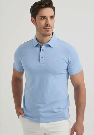 Homme portant un polo bleu clair à manches courtes avec trois boutons, debout les mains dans les poches, regardant légèrement vers la droite.