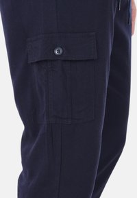 I pantaloni cargo blu marino presentano una tasca laterale con chiusura a bottone, realizzati in un tessuto testurizzato con taglio dritto e una leggera lucentezza.