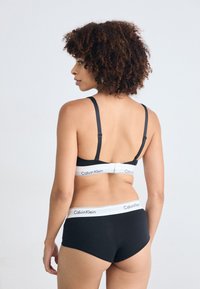 Donna con capelli ricci che indossa un reggiseno nero di Calvin Klein e pantaloni corti abbinati, vista da dietro su uno sfondo chiaro e uniforme.