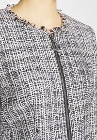 Gros plan d'une veste en tweed noir et blanc avec un col effiloché et une fermeture éclair avant munie d'un anneau rond.