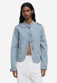 Prugasta traper jakna u svijetloplavoj boji, sa stender ovratnikom, prednjim dugmadi i dva džepa. Glatka tekstura s uskim, krojenim dizajnom.