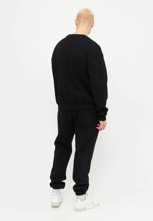 Personne aux cheveux blonds courts, portant un sweat-shirt noir et un pantalon avec des baskets blanches, debout, dos à l'appareil sur un fond blanc uni.