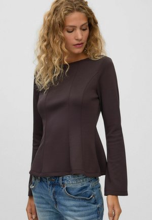 Femme aux cheveux blonds bouclés portant un haut moulant à manches longues foncé et un jean bleu, regardant vers le bas sur un fond uni.