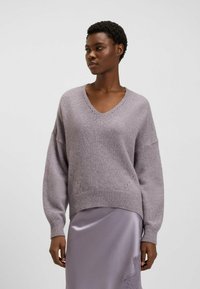 Fioletowy sweter dziany z dekoltem w kształcie litery V, opadającymi ramionami, prążkowanym dołem i subtelnymi szczegółami teksturowymi. Połączony z satynową lawendową spódnicą.