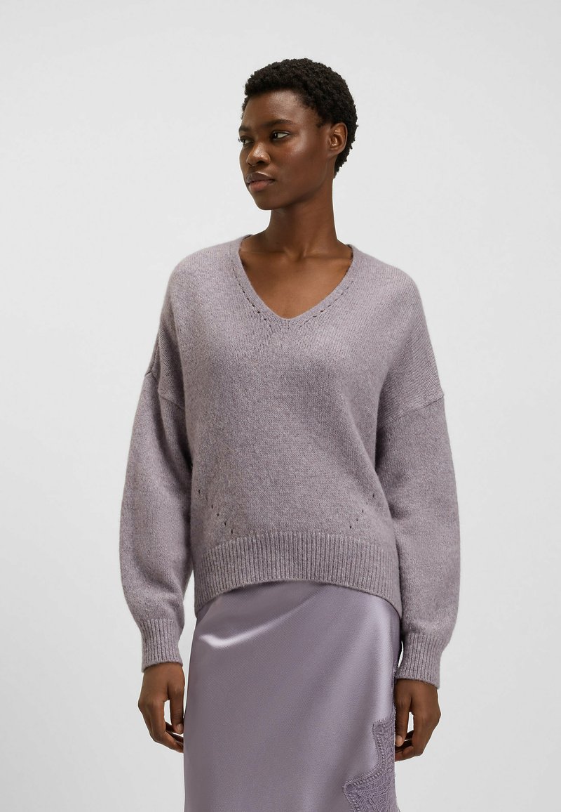 Fioletowy sweter dziany z dekoltem w kształcie litery V, opadającymi ramionami, prążkowanym dołem i subtelnymi szczegółami teksturowymi. Połączony z satynową lawendową spódnicą.