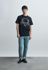 Μαύρο βαμβακερό T-shirt με λευκό λογότυπο και γραφικά Moschino, συνδυασμένο με ανοιχτόχρωμο μπλε τζιν και μαύρα μοκασίνια. Απλό φόντο.